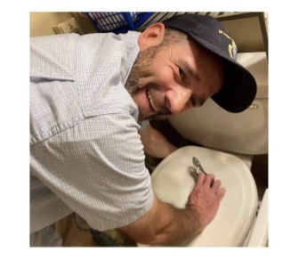 Picture of man leaving over a toilet to fix it wearing a baseball cap and blue collared shirt, smiling. 