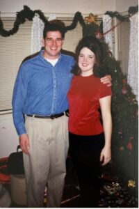 Man in blue collared shirt and kakis with arm around woman wearing red shirt, black pants, standing in front of a Christmas tree. 