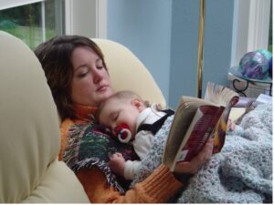 Mother in recliner reading a book while 6-month old child takes a nap on her chest